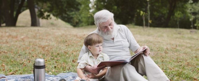 Grand dad reading a book for his grandson during their picnic in their garden. One of the best staycations idea for seniors.